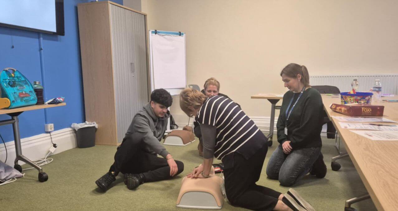 Participants engaging in practical first aid training, demonstrating CPR techniques on manikins in a classroom setting, with instructional materials and a defibrillator visible.