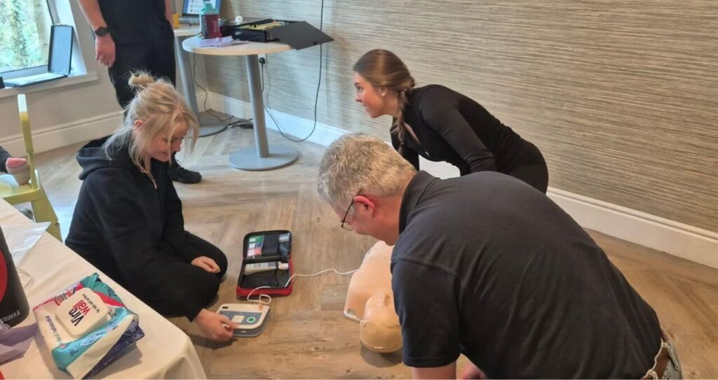 Young woman kneels by a CPR manikin, practicing with an AED trainer device nearby; two adults supervise in the background.
