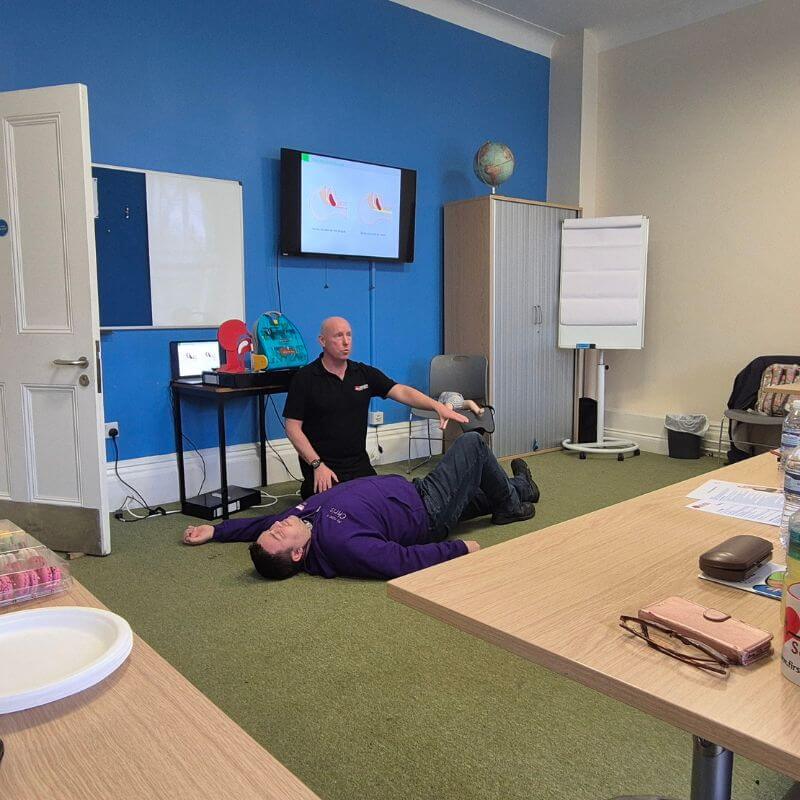 Two men in a training room: one man lying on his back on the carpet while another kneels beside him and points to his leg as if giving a demonstration.
