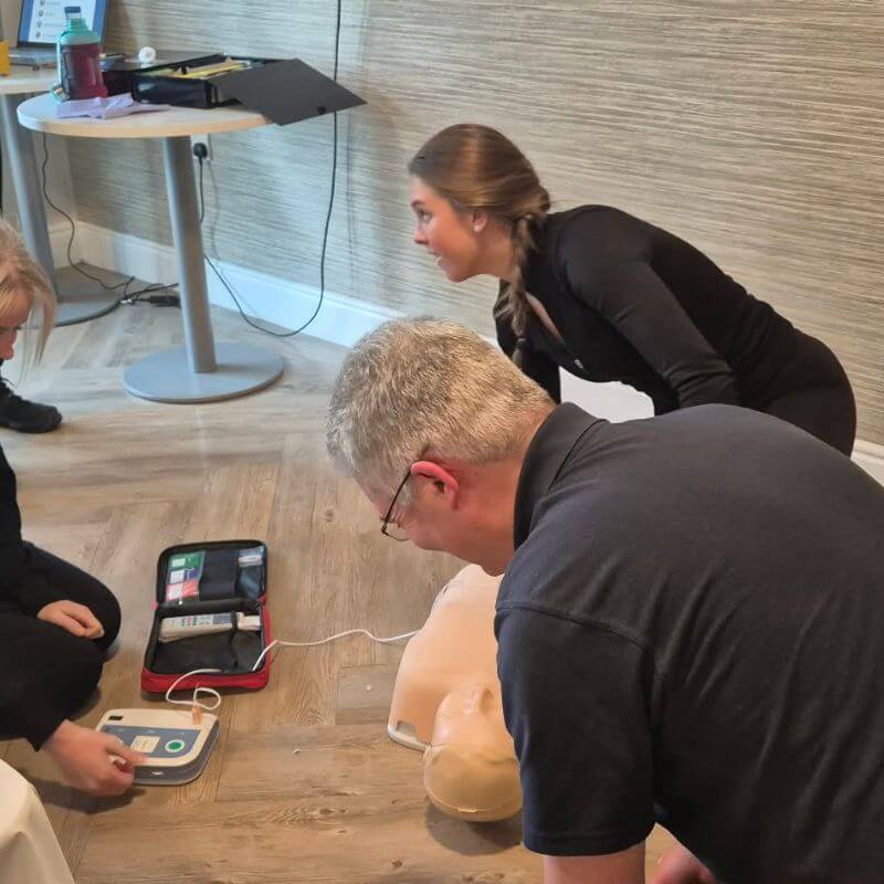 Three people practice CPR on a manikin; an AED trainer and medical kit lie nearby in a training room.