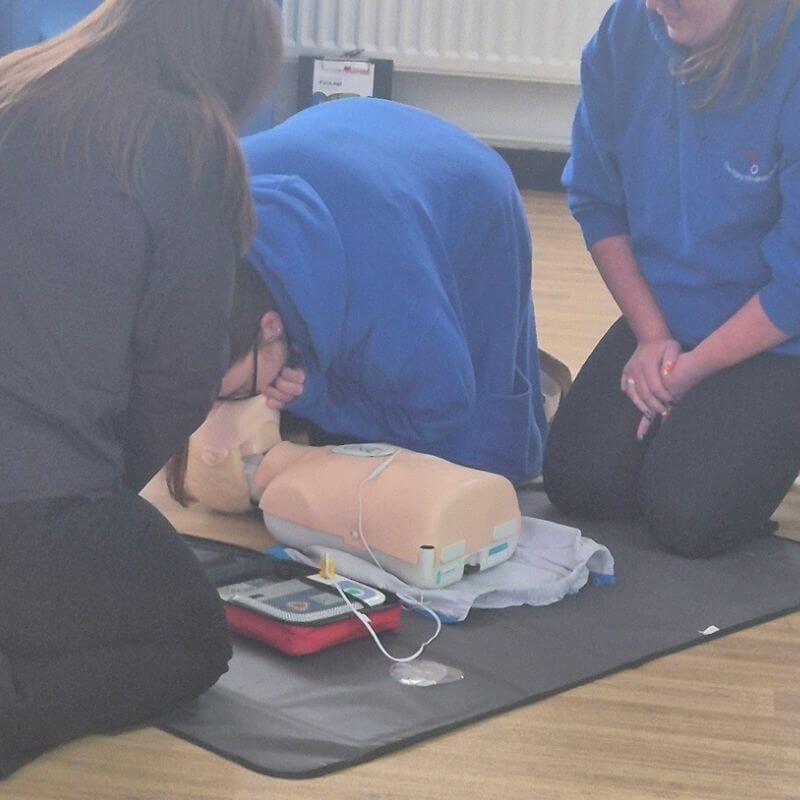 Arrange first aid training in washington | first aid and safety training | first aid and safety training People kneeling around an adult CPR manikin performing training, with an AED trainer and monitor nearby on a mat.