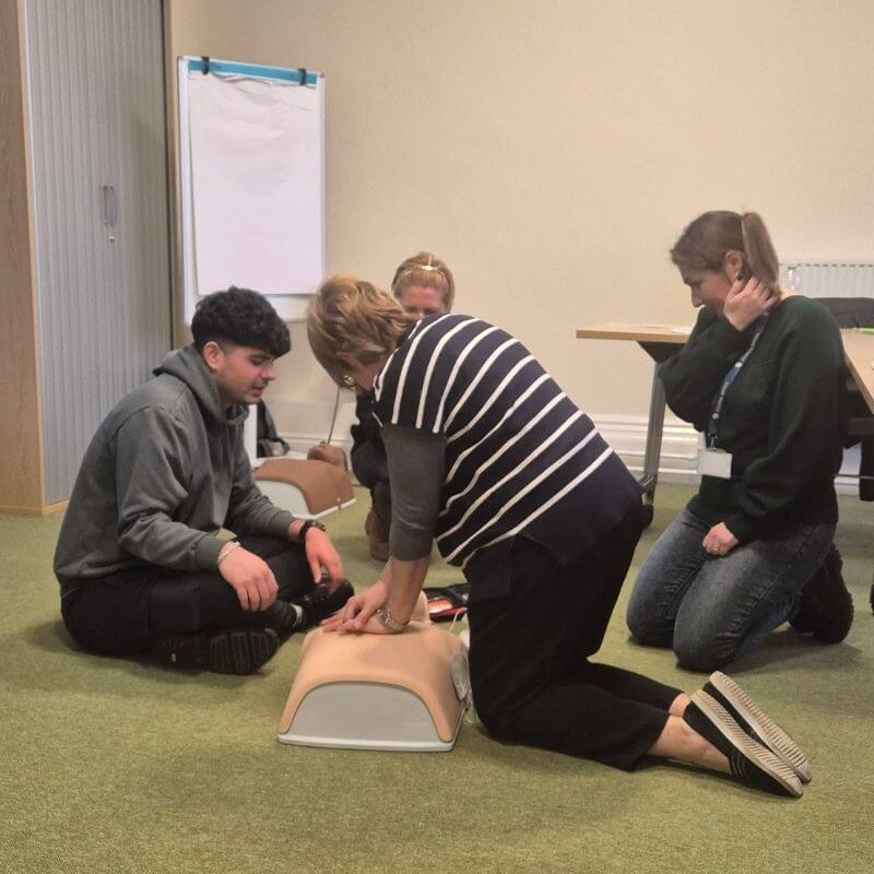 Group of four adults practicing CPR on a life-size manikin in a classroom setting.