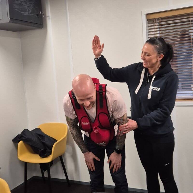 Woman in a dark hoodie high-fives a bald man wearing a red weighted vest during a training session in a small room.