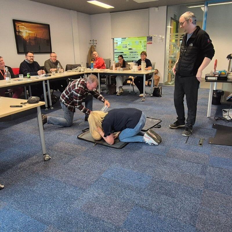 Group of adults in a conference room watching a first-aid demonstration as two participants practice on a person lying on a mat, while others observe nearby.
