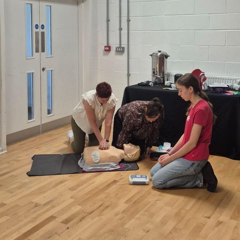 Local first aid training in hebburn | first aid and safety training | first aid and safety training Three people practice CPR on a training manikin: two kneeling and performing chest compressions while a third person watches nearby.