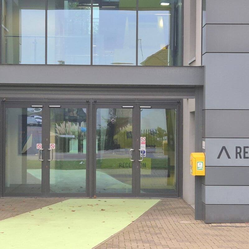 Double glass doors at a modern building entrance, with a yellow intercom on the right and a light green path leading to the doors.