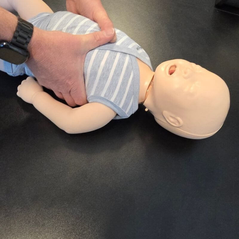 Adult hands performing chest compressions on a baby CPR manikin on a dark table surface.