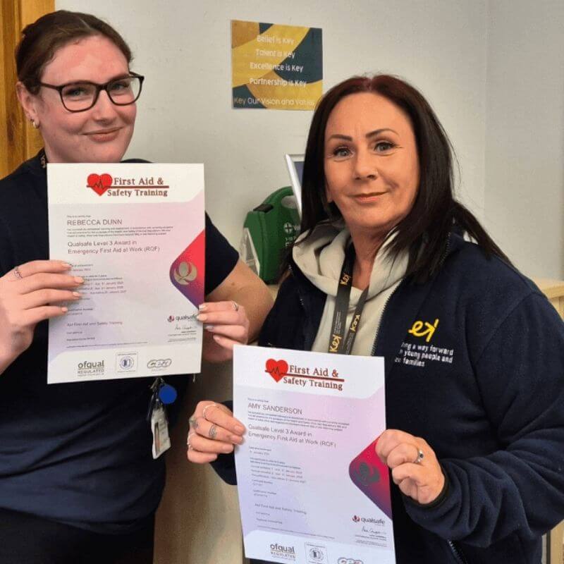 Two women indoors hold up First Aid & Safety Training certificates, smiling at the camera with lanyards visible.
