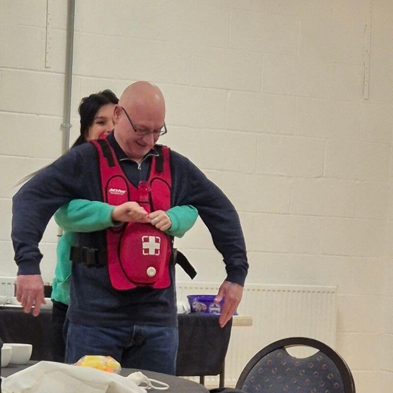 We deliver first aid training across the north east | first aid and safety training | first aid and safety training Man wearing a red first-aid vest with a white cross, smiling as someone helps adjust it indoors during a training session.