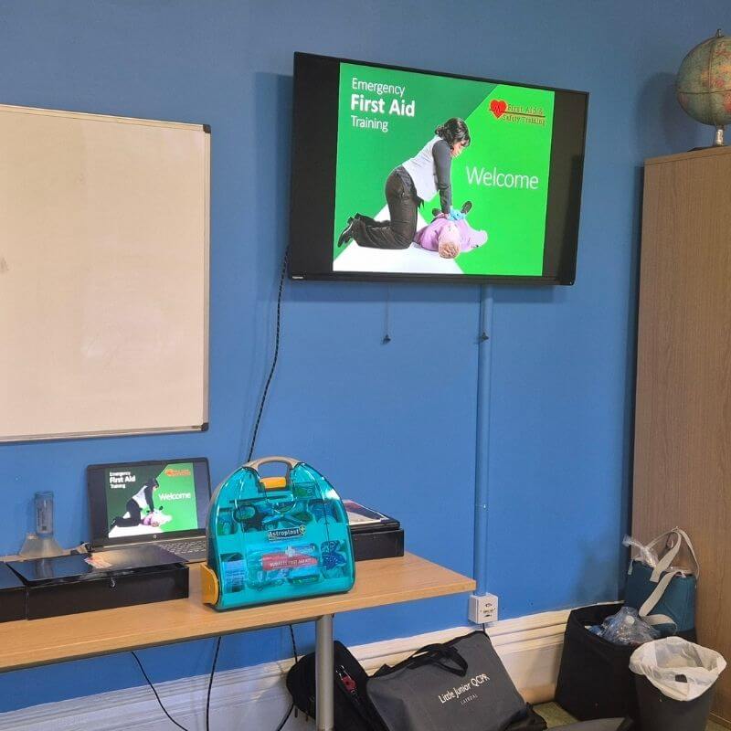 TV screen shows 'Emergency First Aid Training' with a trainer performing chest compressions beside a mannequin in a classroom.