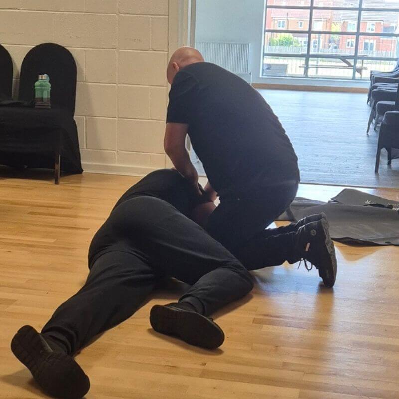 Two men on a wooden floor; one kneels over a person lying face down, likely giving first aid in a training room with windows in the background.