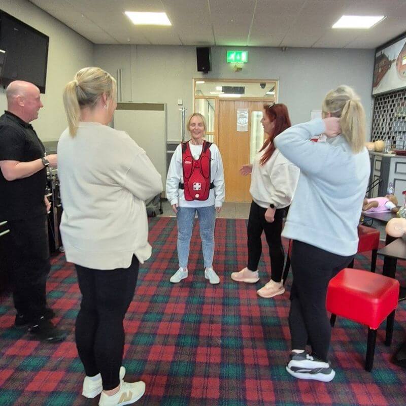 A group of five adults standing in a circle in a training room, with a woman in the center wearing a red first-aid vest and blue jeans leading the session.