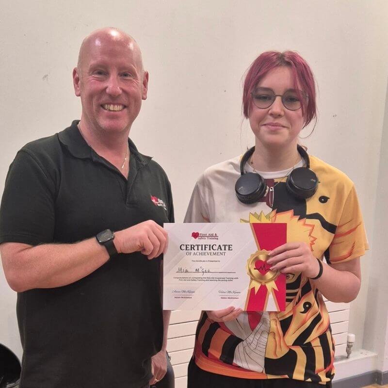 A man in a black polo stands beside a young woman with red hair holding a certificate of achievement and a decorative ribbon while smiling at the camera in a plain room.