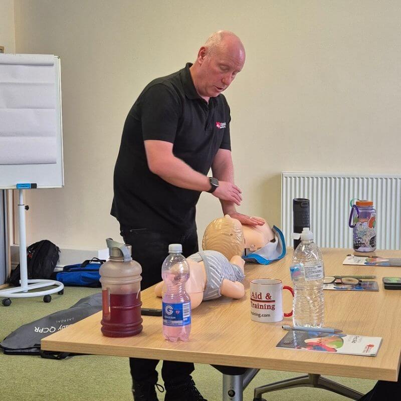 Instructor demonstrates chest compressions on a CPR manikin during a first-aid training session at a classroom table.