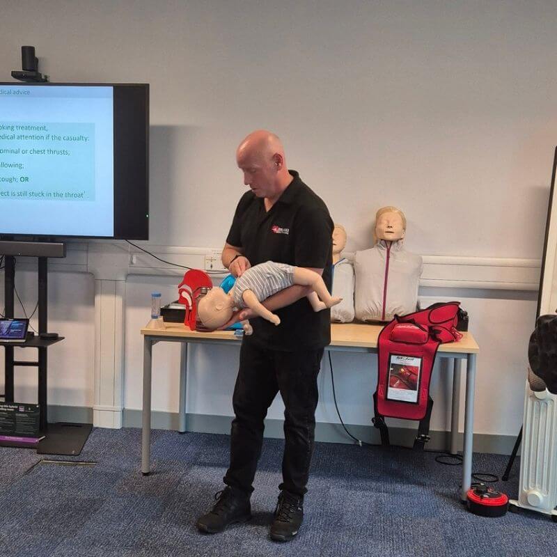 Instructor demonstrates infant CPR on a lifelike baby manikin in a training room with mannequins and teaching screen nearby