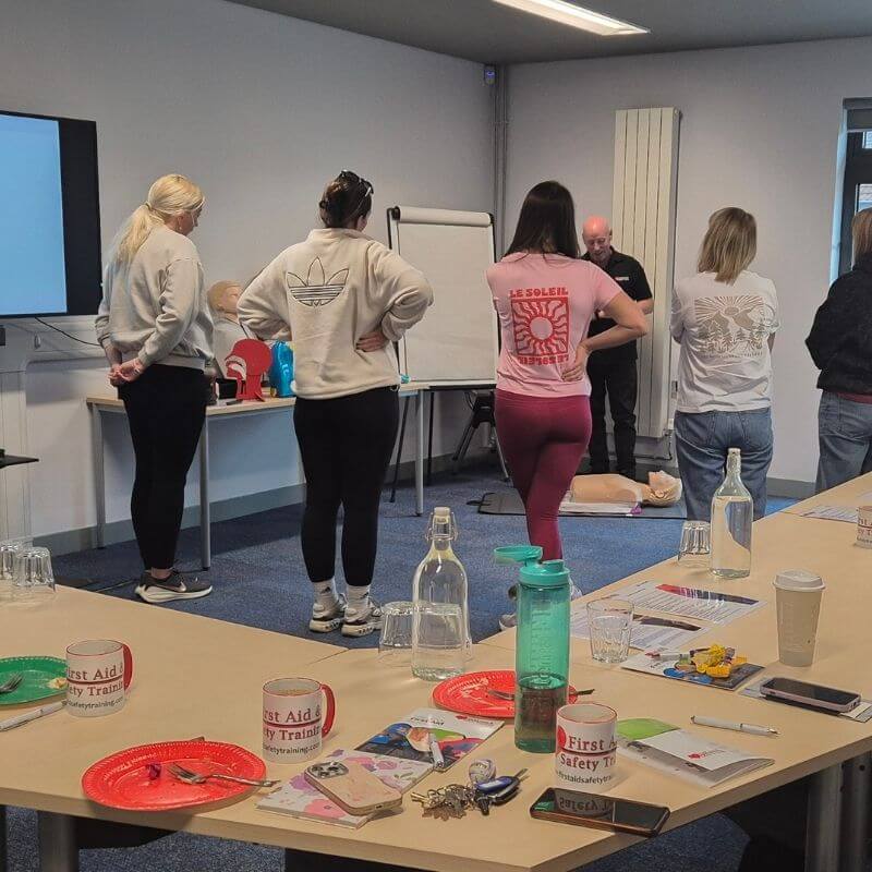Group of adults in a training room gathered around CPR mannequins as an instructor leads a first-aid class by a flip chart.