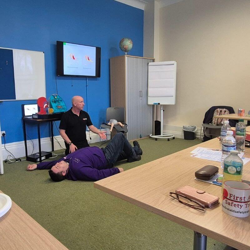 CPR training scene: instructor kneeling beside a person lying on the floor as a presentation screen and classroom equipment are visible in a blue-walled room.