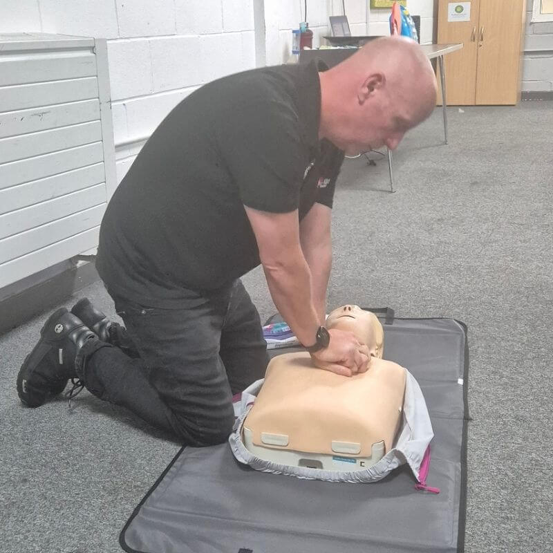 Man kneeling on the floor and performing chest compressions on an adult CPR dummy during training.