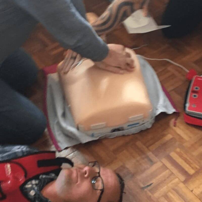 Person performing chest compressions on a CPR training manikin on a wooden floor; a red AED trainer lies nearby.