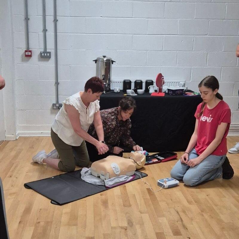 Two women and a young girl practice CPR on a manikin on a gym floor, with a refreshment table and equipment in the background.