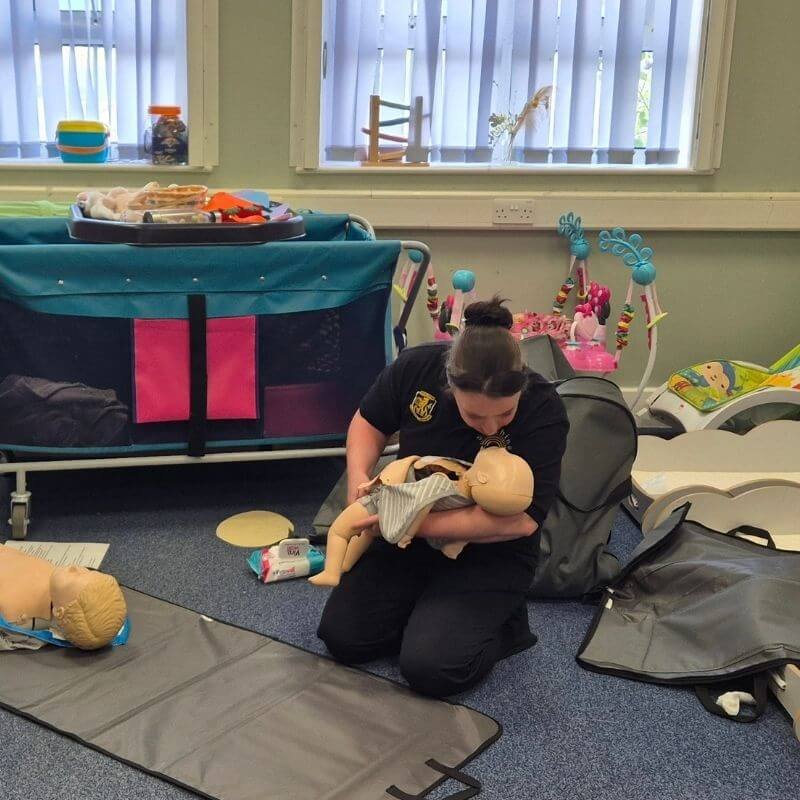 Caregiver in uniform practicing infant CPR on a baby mannequin in a nursery training room with cribs and toys nearby